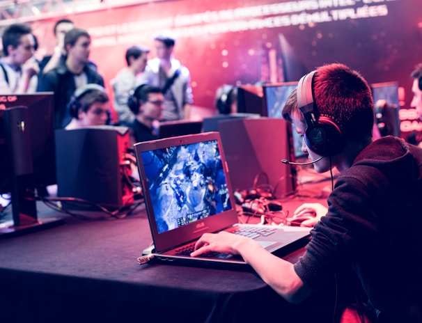 Several people are seated and focused on computer screens, wearing headsets, in an environment that appears to be a gaming tournament or esports event. The room is dimly lit with neon lighting, highlighting screens and creating an intense atmosphere.