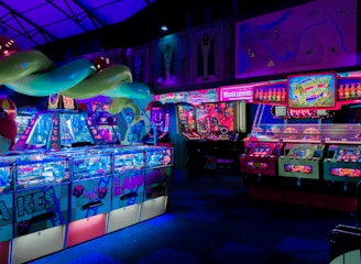 An indoor arcade filled with vibrant gaming machines lit with neon blue and pink lights. The scene includes various arcade games with flashing screens and colorful displays, and a large overhead structure resembling a green snake. The background shows illuminated signs and themed decor, contributing to an immersive entertainment atmosphere.