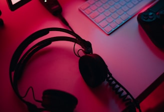 A dimly lit gaming setup with a monitor displaying a game's menu on the screen. A mechanical keyboard sits in front of the monitor. A pair of over-ear headphones and a game controller rest on the desk. The scene is bathed in a red and blue light, creating a moody gaming atmosphere.