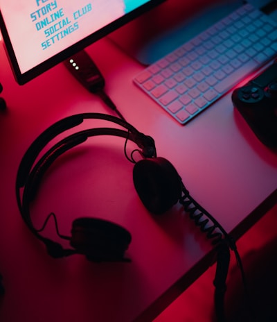 A dimly lit gaming setup with a monitor displaying a game's menu on the screen. A mechanical keyboard sits in front of the monitor. A pair of over-ear headphones and a game controller rest on the desk. The scene is bathed in a red and blue light, creating a moody gaming atmosphere.