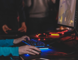 A person is engaged in a video game at a computer setup, using a keyboard and mouse. The scene is dimly lit, with the glow from the monitor reflecting off the hands of the player. Several other people are present in the background, indicating a social or competitive gaming environment.