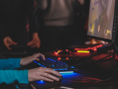 A person is engaged in a video game at a computer setup, using a keyboard and mouse. The scene is dimly lit, with the glow from the monitor reflecting off the hands of the player. Several other people are present in the background, indicating a social or competitive gaming environment.