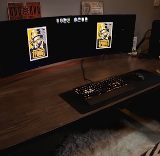 A gaming setup featuring a large curved monitor displaying PUBG logos on the screens. A keyboard and mouse are positioned on a large desk pad. A pair of headphones rests on the desk alongside a mobile phone. A drink in a Starbucks cup is placed to the right, while a desktop tower is seen in the background on the right side.