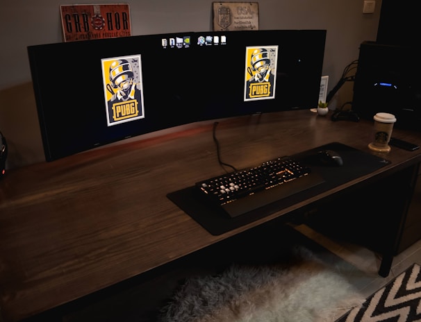 A gaming setup featuring a large curved monitor displaying PUBG logos on the screens. A keyboard and mouse are positioned on a large desk pad. A pair of headphones rests on the desk alongside a mobile phone. A drink in a Starbucks cup is placed to the right, while a desktop tower is seen in the background on the right side.