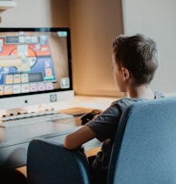 A young person is sitting on a blue chair at a desk, focused on a computer screen displaying a colorful card game. The scene appears to be set in a cozy, well-lit room.