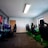 A well-lit gaming room features multiple gaming stations with high-backed chairs and monitors. The room has a minimalist setup with green and black color tones. Large posters adorn the walls, and the room appears clean and organized.