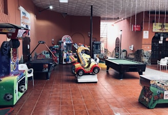 An indoor arcade area featuring various gaming machines, including an air hockey table and a ride-on toy car. The room has a tiled floor and several colorful gaming devices scattered around. Chairs and arcade signage are visible, creating a playful and entertainment-focused atmosphere.