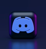A 3D-style block with a blue logo in the center, set against a dark background. The logo resembles a game controller with two rounded edges and two circles on the front. The block has a subtle gradient, transitioning from dark to purple on its sides.
