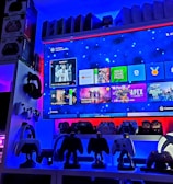 A modern gaming setup featuring a large wall-mounted screen displaying a gaming console interface. The room is decorated with gaming-themed items such as controllers and collectibles. The ambient lighting includes shades of blue and red, creating a vibrant atmosphere.