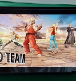 A handheld gaming console displays a screenshot of a fighting game featuring cartoonish characters in dynamic poses. The characters are standing in a virtual arena setting with a sky and cloud backdrop. The console is placed on a green surface.