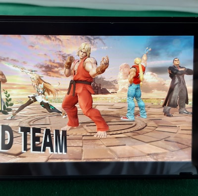 A handheld gaming console displays a screenshot of a fighting game featuring cartoonish characters in dynamic poses. The characters are standing in a virtual arena setting with a sky and cloud backdrop. The console is placed on a green surface.