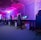 A row of gaming computers with large monitors and racing-style chairs is set up along the interior of a modern room. The space is illuminated with vibrant purple and blue LED lighting, creating a futuristic atmosphere. The room is organized and clean, suggesting a professional gaming or esports environment.