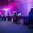 A row of gaming computers with large monitors and racing-style chairs is set up along the interior of a modern room. The space is illuminated with vibrant purple and blue LED lighting, creating a futuristic atmosphere. The room is organized and clean, suggesting a professional gaming or esports environment.