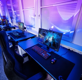 A row of modern gaming setups is arranged neatly along a window-lined wall. Each station includes a high-resolution monitor displaying vivid graphics, a mechanical keyboard with colored lighting, a gaming mouse, and an ergonomic chair. The room is illuminated with ambient blue and purple lights, creating a sleek and futuristic atmosphere.