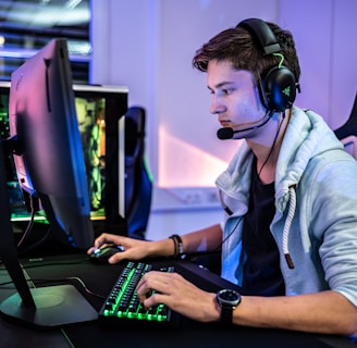 A young person is intensely focused on a computer screen while wearing a headset with a microphone. They are using a gaming keyboard and mouse, sitting in a gaming chair. The room has cool lighting, with a visible PC tower.