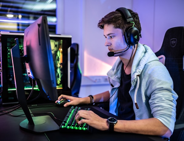 A young person is intensely focused on a computer screen while wearing a headset with a microphone. They are using a gaming keyboard and mouse, sitting in a gaming chair. The room has cool lighting, with a visible PC tower.