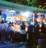 A group of people standing in front of an illuminated amusement arcade, engaging with various gaming machines. The environment is bustling with activity, and colorful lights create a vibrant atmosphere. The ceiling is decorated with large, playful images of dice and other game-related graphics, enhancing the lively mood.