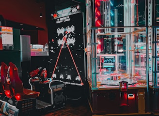 An arcade setting featuring a classic Space Invaders game setup and a modern gaming machine with seats. Bright neon lights illuminate the area, with scores displayed on digital screens. A bright and visually engaging environment full of electronic components.