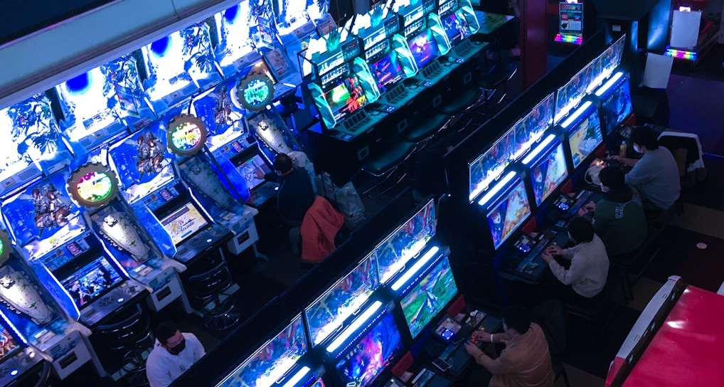 A bustling arcade filled with rows of gaming machines, each with vibrant displays and controls. Several people are seated at these machines, deeply engaged in playing games. Bright fluorescent lights cast a blue and neon glow across the room, enhancing the lively and energetic atmosphere.