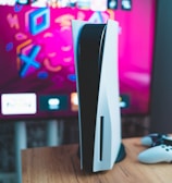A gaming console is placed on a wooden table with a game controller nearby. In the background, a television screen displays colorful graphics, including geometric shapes and vibrant colors.
