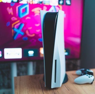 A gaming console is placed on a wooden table with a game controller nearby. In the background, a television screen displays colorful graphics, including geometric shapes and vibrant colors.