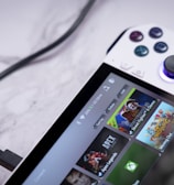A handheld gaming console with a white body and colorful buttons features a screen displaying various game icons. The device is connected to a power source via a black cable and rests on a marble surface. The interface shows games like Apex and Street Fighter.