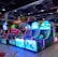 A brightly lit arcade filled with colorful gaming machines sits under ceiling lights. Neon lights illuminate various game stations, creating a playful and vibrant environment. A few red chairs are scattered around the reflective, tiled floor, and a person is observing one of the machines. The atmosphere is lively and cheerful, featuring games with cartoonish designs and engaging themes.