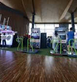 A vibrant arcade setting with several gaming machines lined up, including dance and rhythm game setups. People are engaged with the games, absorbed in their activities. The flooring is wooden, and the ceiling has a geometric design. Natural light filters through large windows, creating a lively atmosphere.