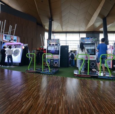 A vibrant arcade setting with several gaming machines lined up, including dance and rhythm game setups. People are engaged with the games, absorbed in their activities. The flooring is wooden, and the ceiling has a geometric design. Natural light filters through large windows, creating a lively atmosphere.