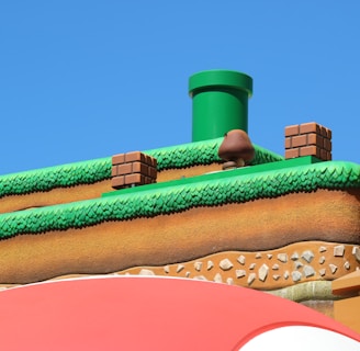 A colorful, stylized scene depicting a platform from a video game setting. The scene features a grass-topped surface with soil layers beneath, a green pipe, brick blocks, and a character resembling a mushroom. The sky is clear and blue, creating a vibrant backdrop.