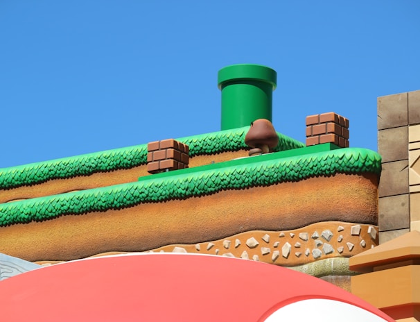 A colorful, stylized scene depicting a platform from a video game setting. The scene features a grass-topped surface with soil layers beneath, a green pipe, brick blocks, and a character resembling a mushroom. The sky is clear and blue, creating a vibrant backdrop.