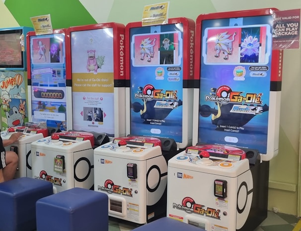 Several arcade gaming machines with multiple screens displaying Pokémon-themed content are positioned in a colorful arcade setting. There are people interacting with the machines; some are seated, and others are standing. The area features different gaming options, including a jumping game with bright, inviting graphics.